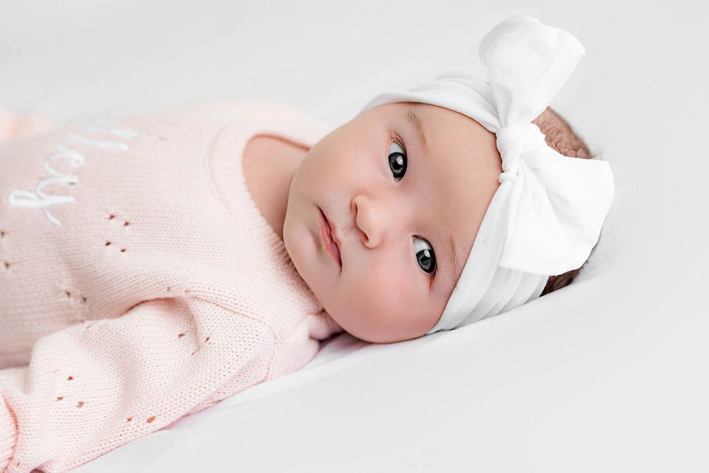 Newborn baby girl in pink knit outfit and white bow hat lying peacefully on a white background.