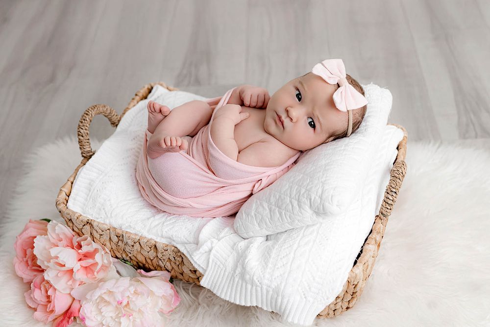 Newborn baby girl wrapped in pink swaddle lying in wicker basket with white blanket and pink flowers.