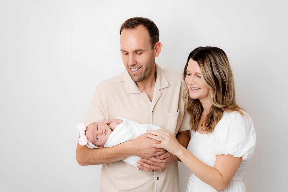A couple smiles lovingly at their newborn baby wrapped in white against a clean white studio background.