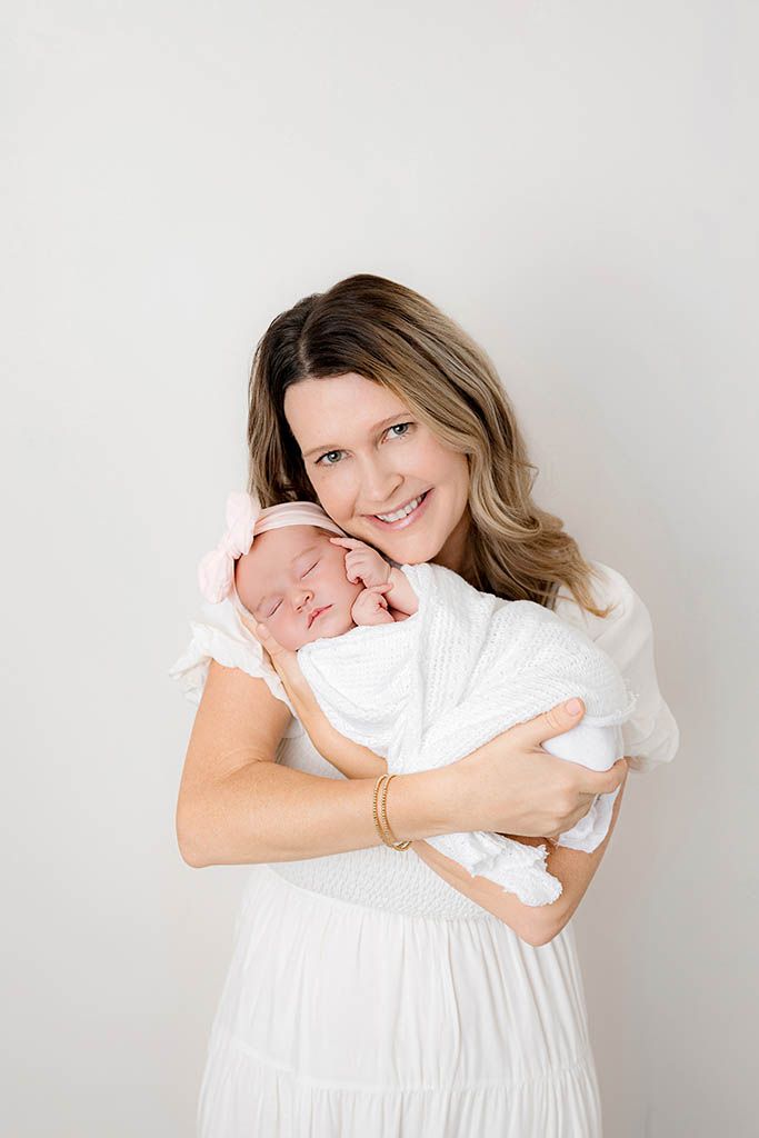 Smiling mother in white dress tenderly holding her swaddled newborn baby girl with pink bow.