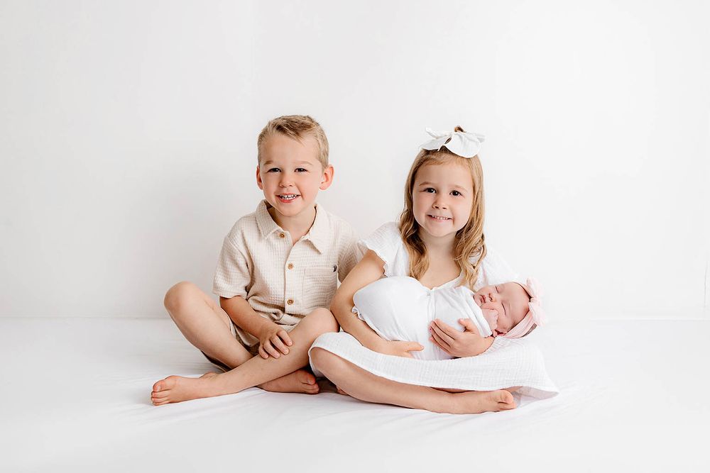 Two older siblings smile while holding their swaddled newborn baby sister during a professional white studio photo session.