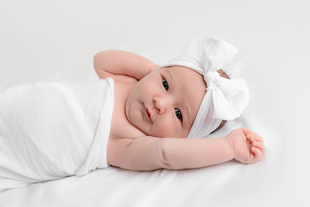 Newborn baby girl swaddled in white wrap wearing a white bow headband, lying on a white background.