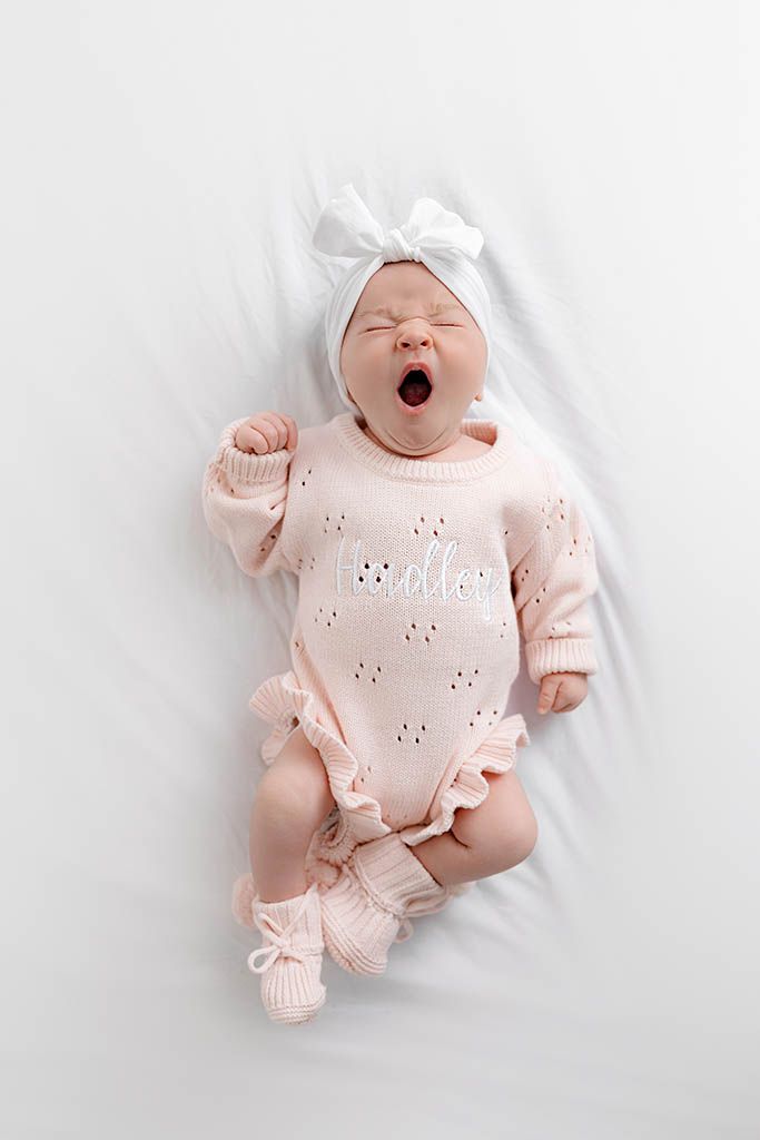 Newborn baby yawning in pink knit onesie with white bow headband and booties, lying on white blanket.