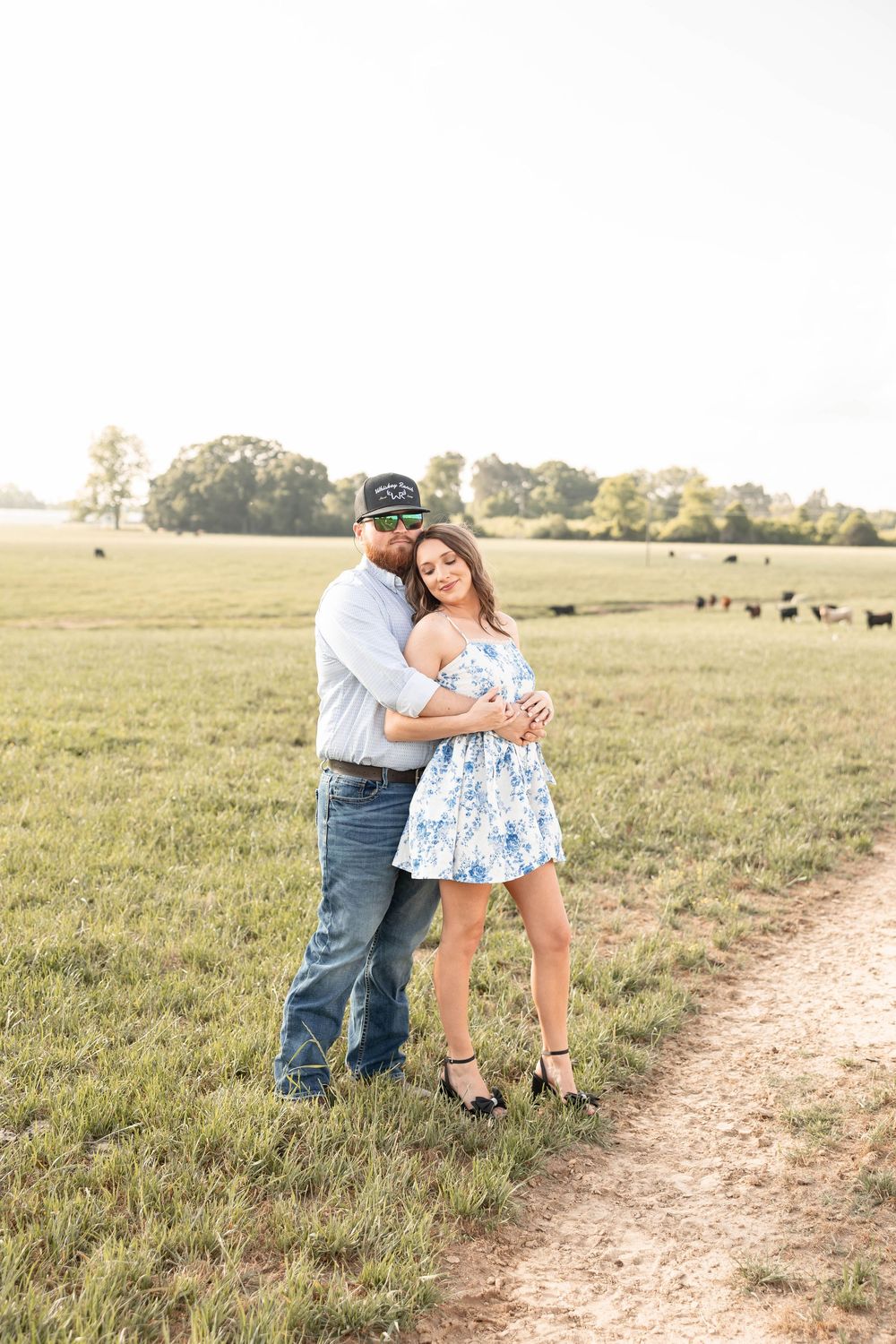 Couple embracing in a sunlit green pasture, woman in floral dress and man in jeans and cap, cattle grazing in background.