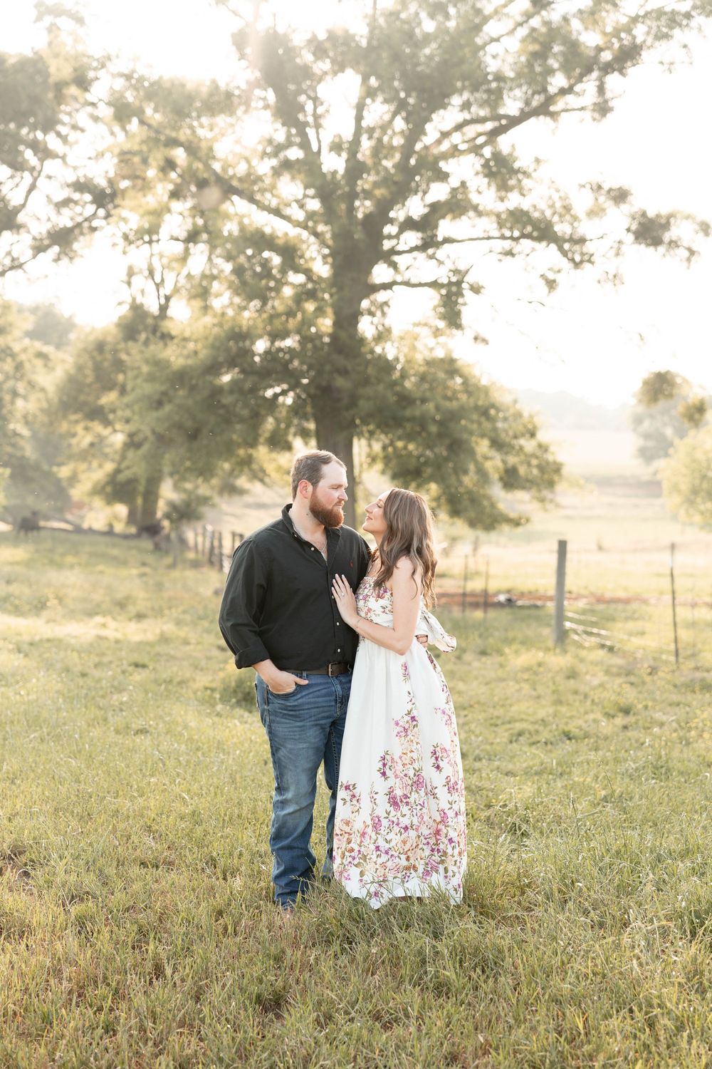 Couple sharing a romantic moment in a golden hour field, man in black shirt and woman in floral maxi dress near a large oak tree.