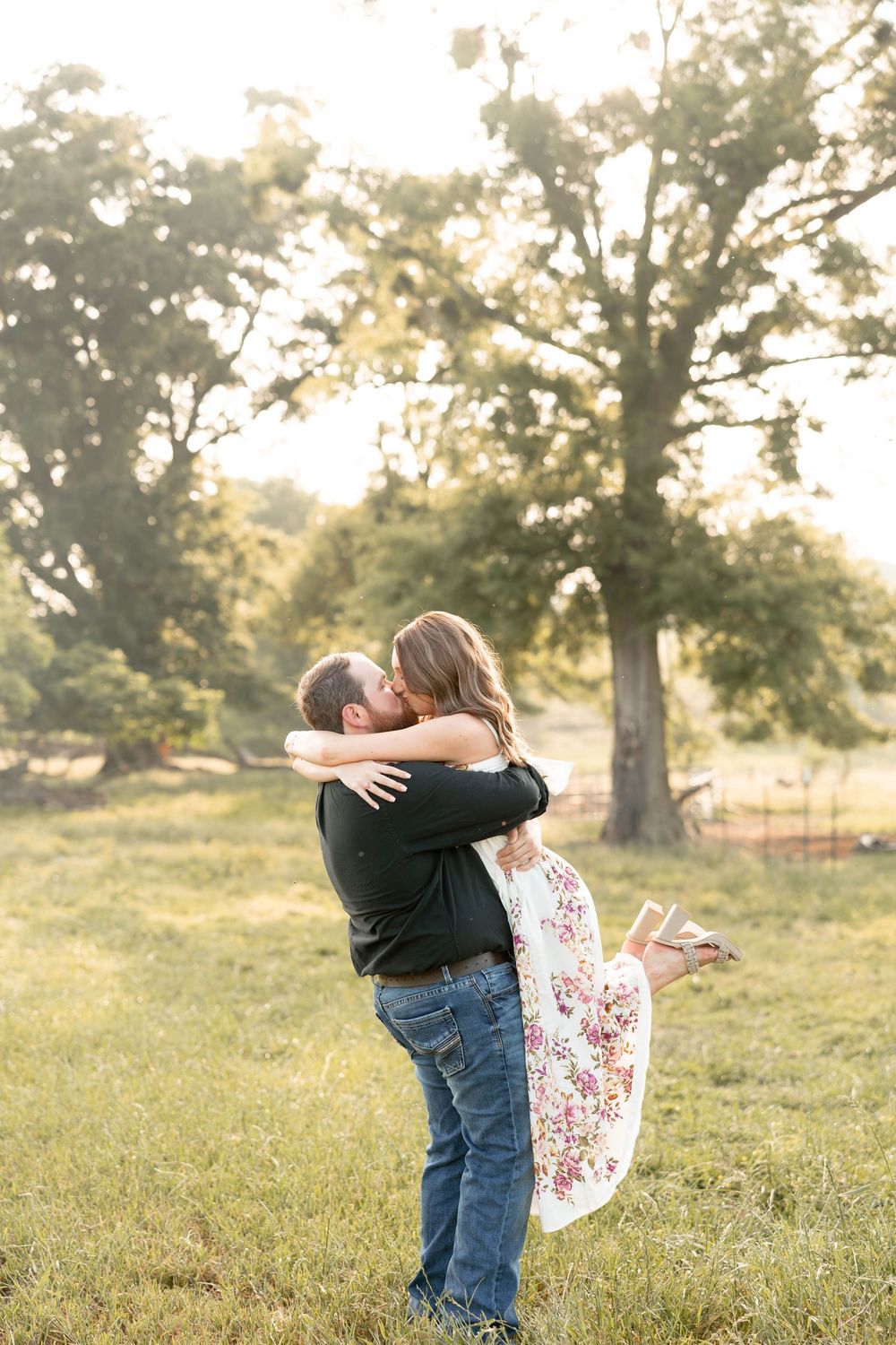 Man lifting woman in floral dress for a kiss in a sunlit green field with large trees in the background.