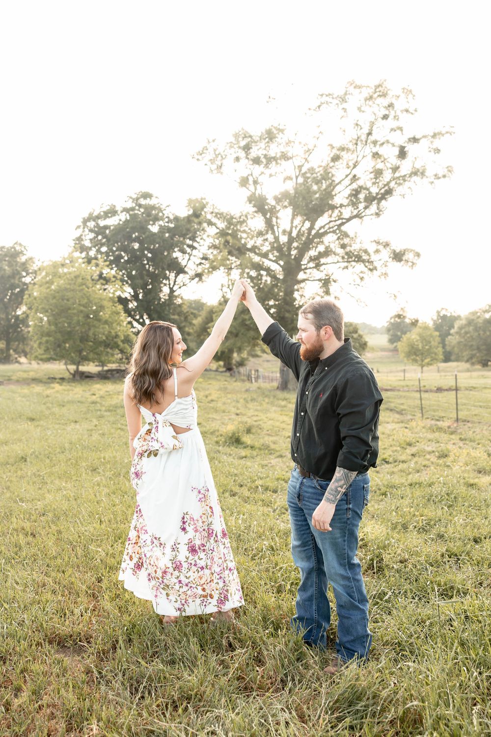 Couple dancing in a sunlit green field, woman in floral dress and man in black shirt holding hands raised.