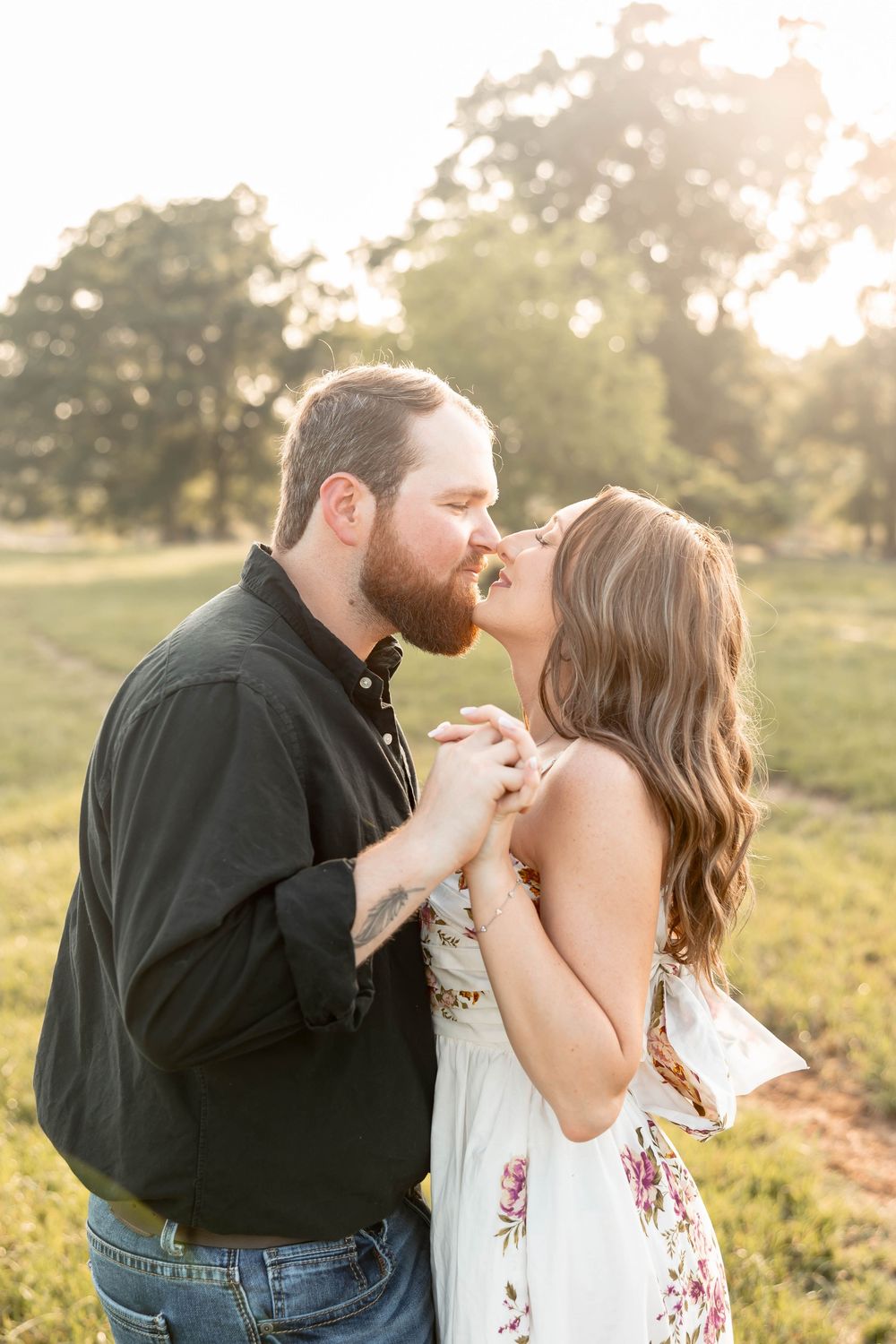 Couple sharing a tender kiss at sunset in a green field, man in black shirt holding hands with woman in white floral dress.