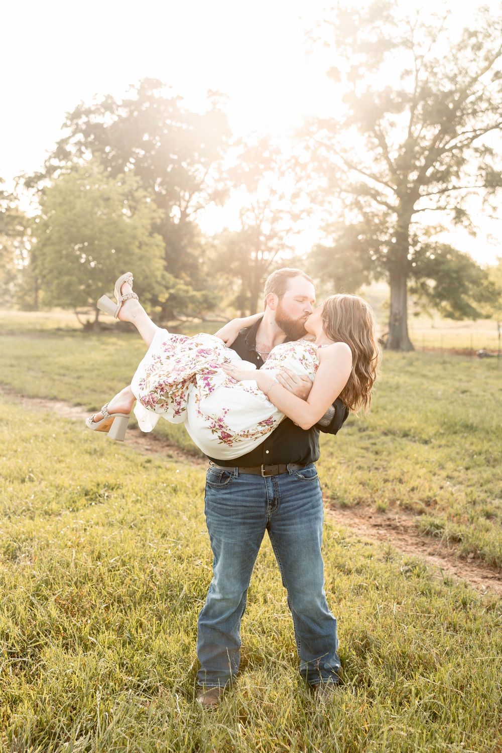 Man in jeans lifts woman in floral dress for a kiss in a sunlit green field with trees in the background.