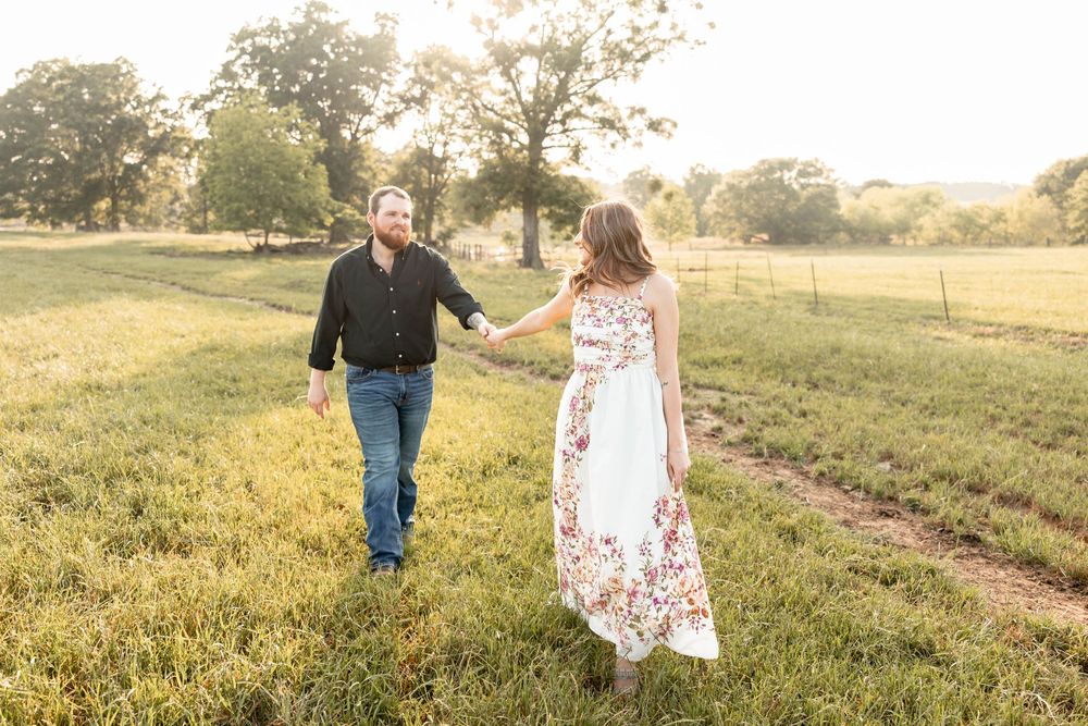 Couple walking hand in hand through a lush green field at golden hour, woman in floral maxi dress.