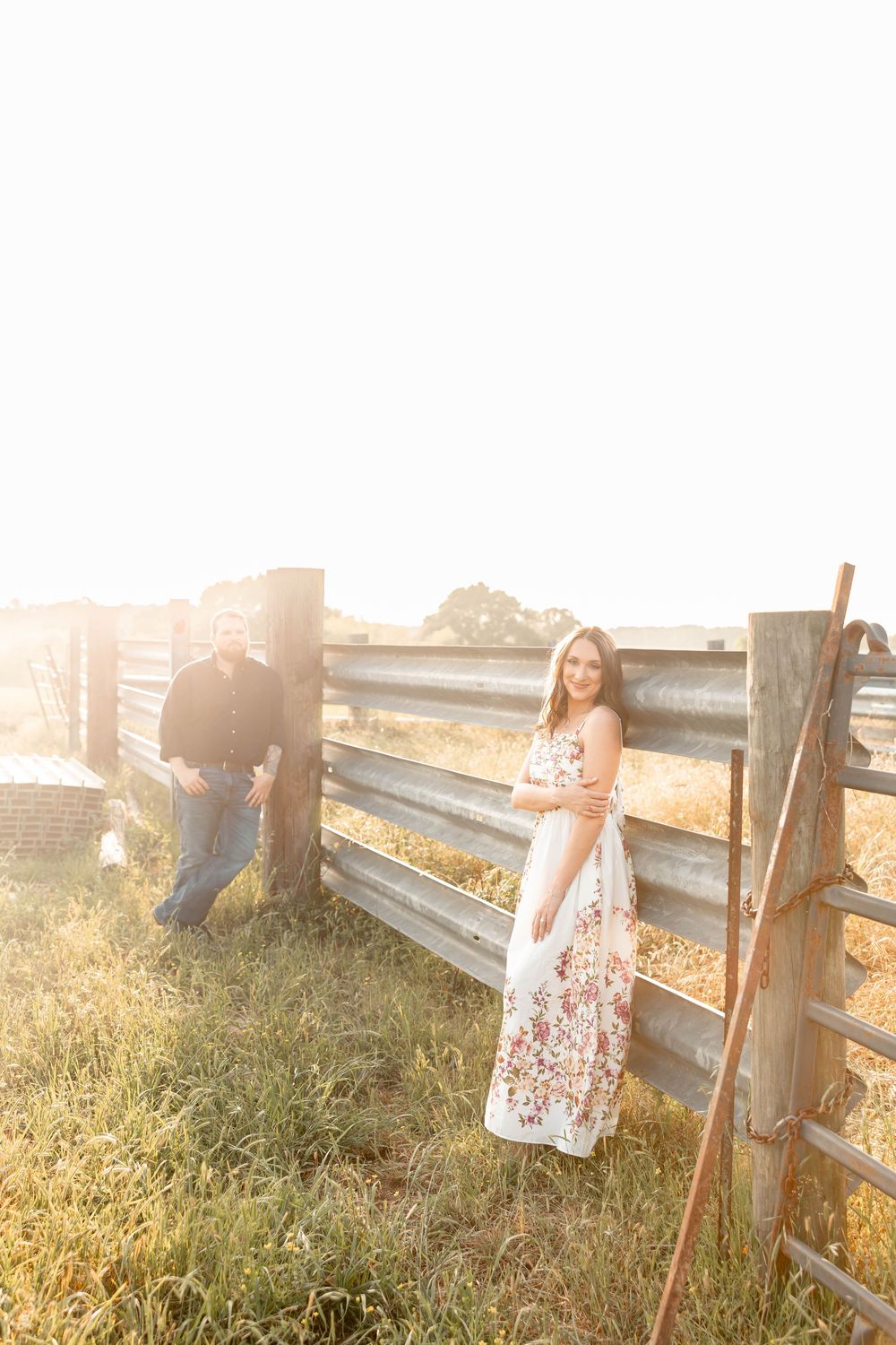 Couple posing by a rustic wooden fence in a sunlit field, woman wearing a white floral maxi dress.