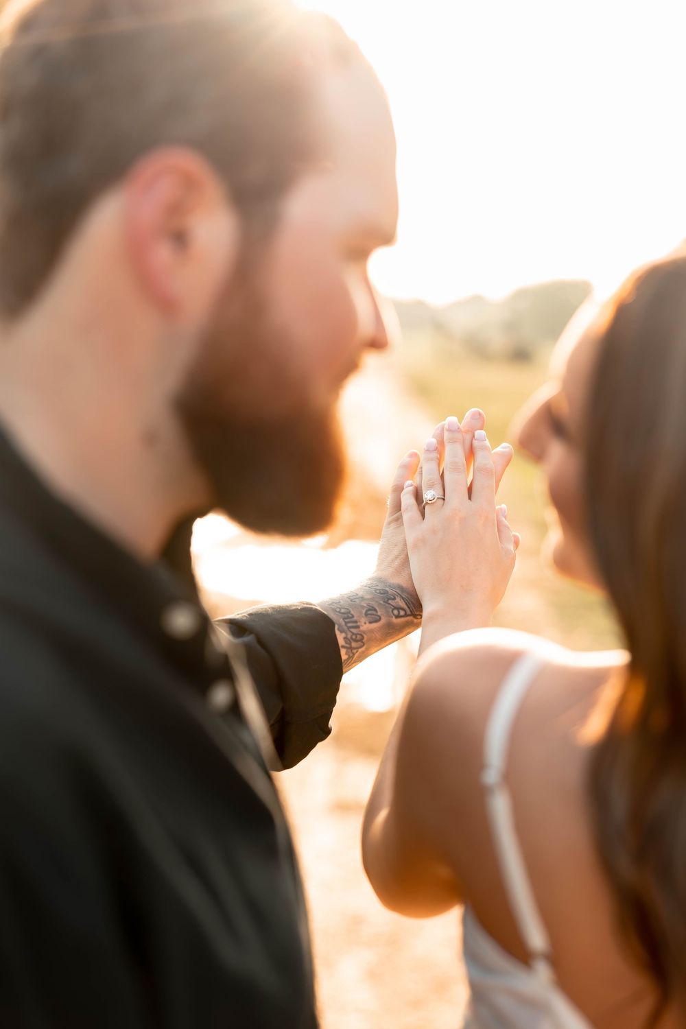 Couple holding hands showing engagement ring with tattoos and golden sunset bokeh background.