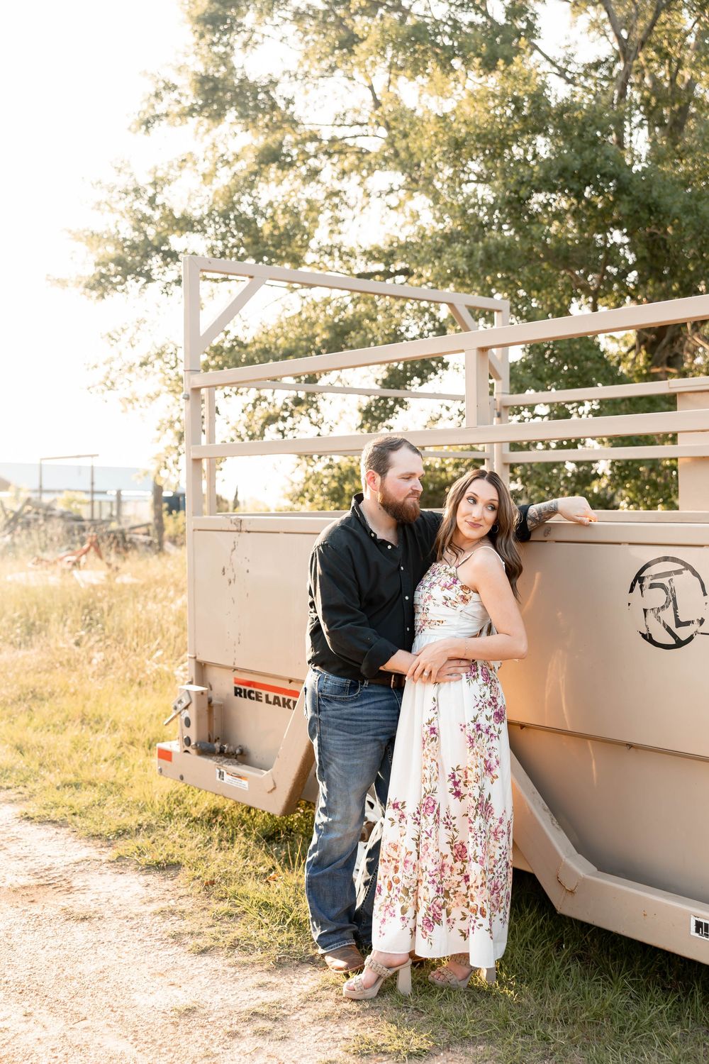 Couple leaning against a tan livestock trailer in a golden hour field, woman in white floral dress.