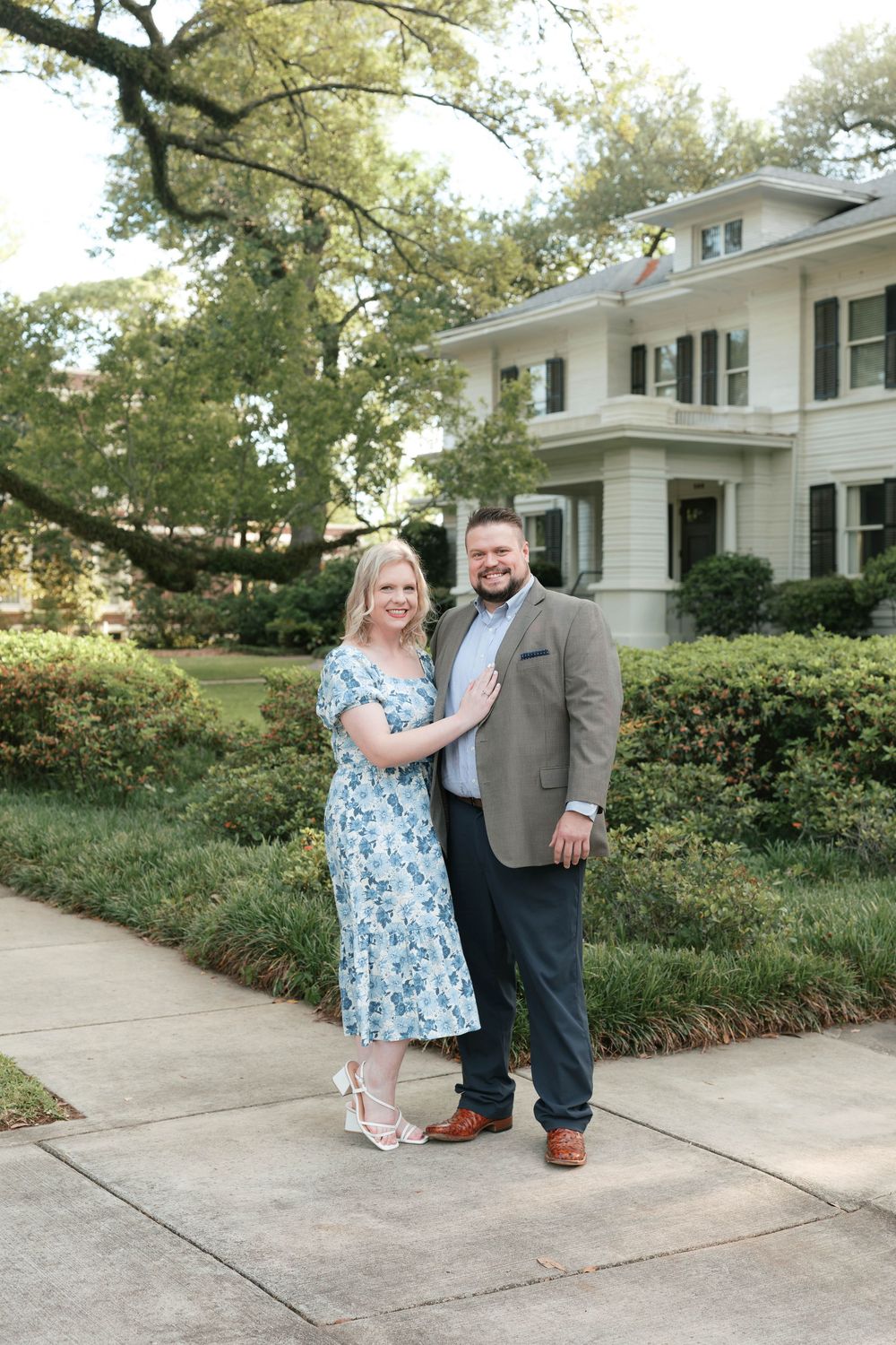 Couple posing on a sidewalk in front of a white colonial home, woman in floral dress, man in tan blazer.