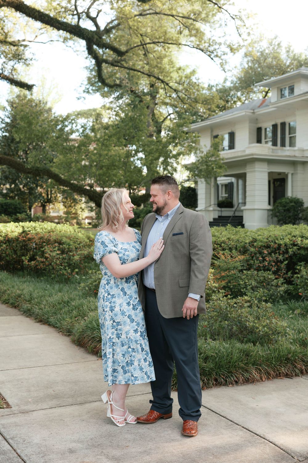 Couple in floral dress and gray blazer smiling at each other on a sidewalk near a white house.