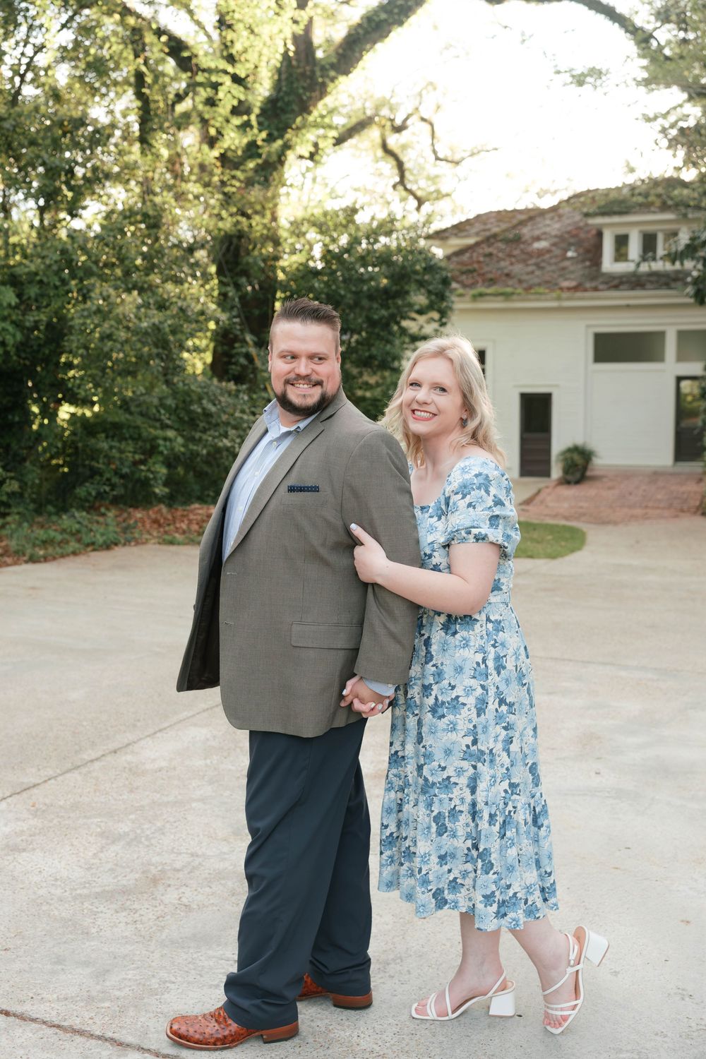 A smiling couple poses outdoors, man in gray blazer and woman in blue floral dress, holding hands near a white building.