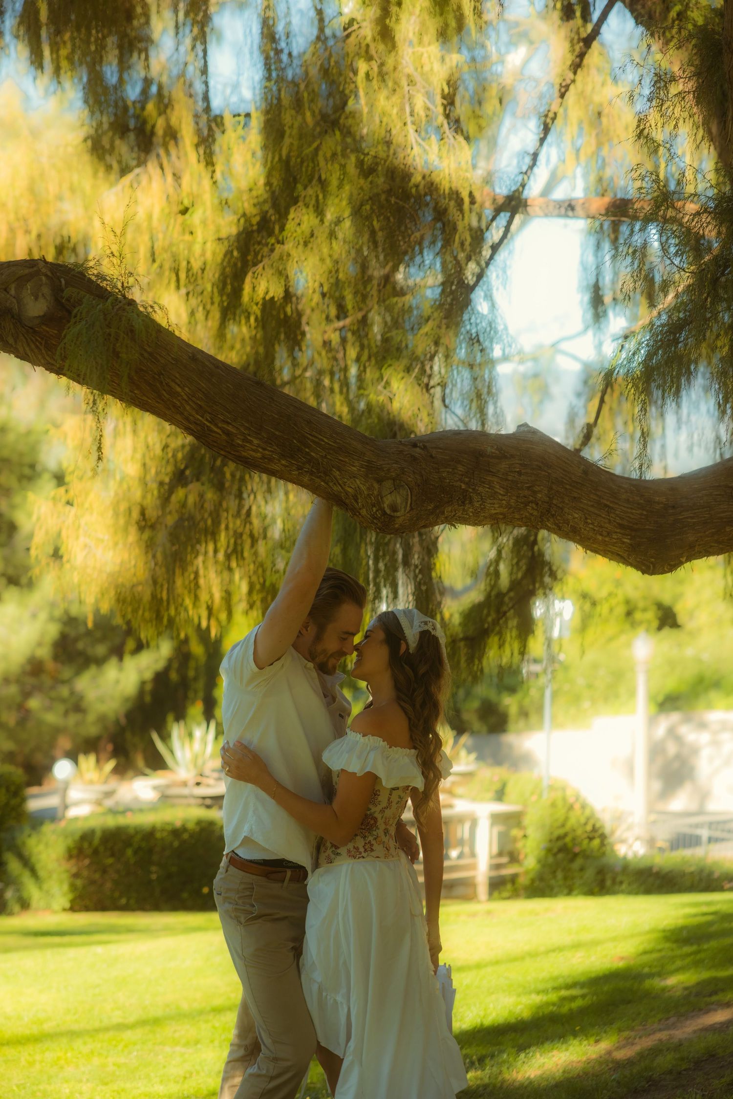Romantic couple embracing under a large tree at the Kimberly Crest House & Gardens, woman in white dress, man in casual attire.