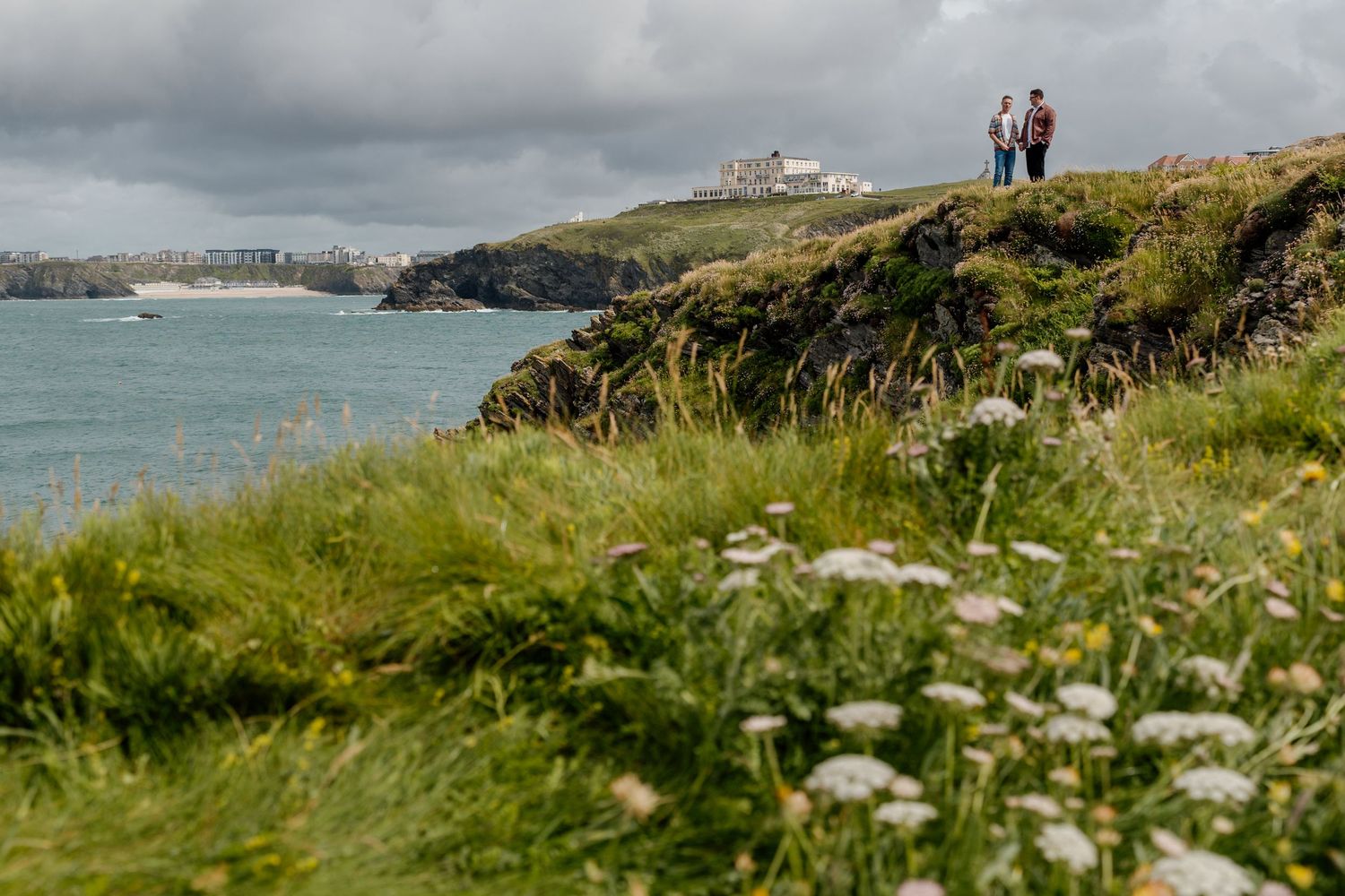 Two people walk along a dramatic coastal cliff path with wildflowers, overlooking a turquoise bay and headland.