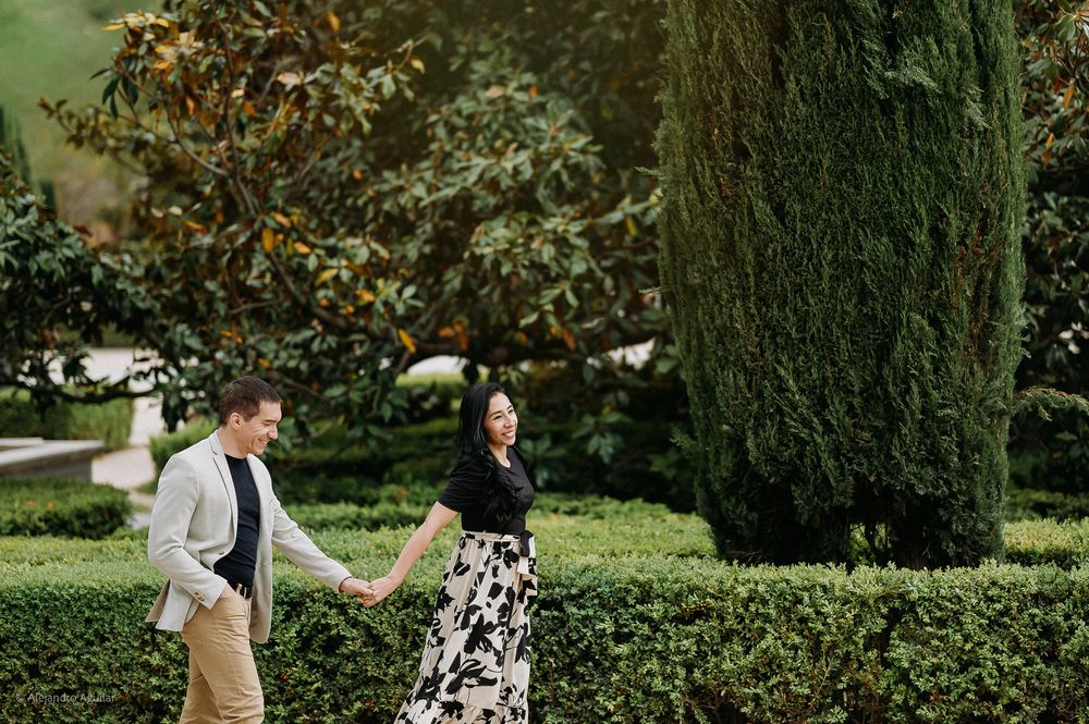 Couple strolling hand in hand through a lush green garden with tall cypress trees.