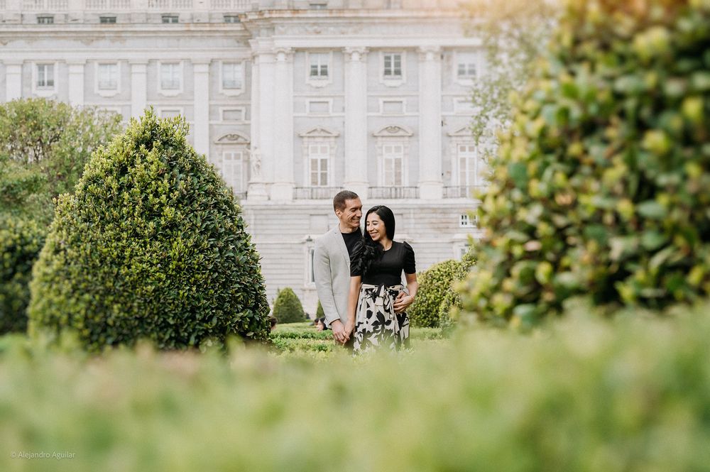 Couple sharing a tender moment among lush green hedges near a grand palace.
