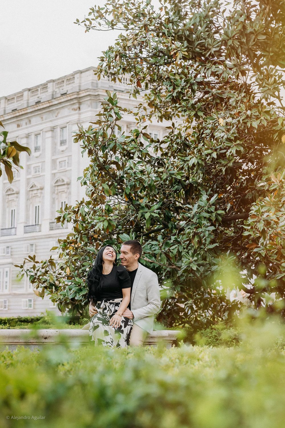 Couple sitting and laughing together in a garden with a white palace backdrop.
