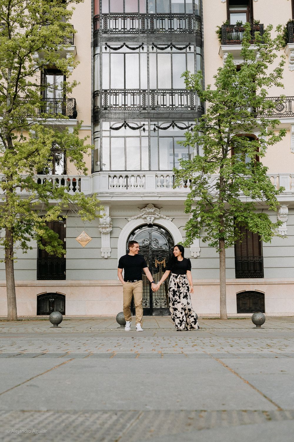 Couple holding hands and smiling before an elegant Art Nouveau building in Madrid.