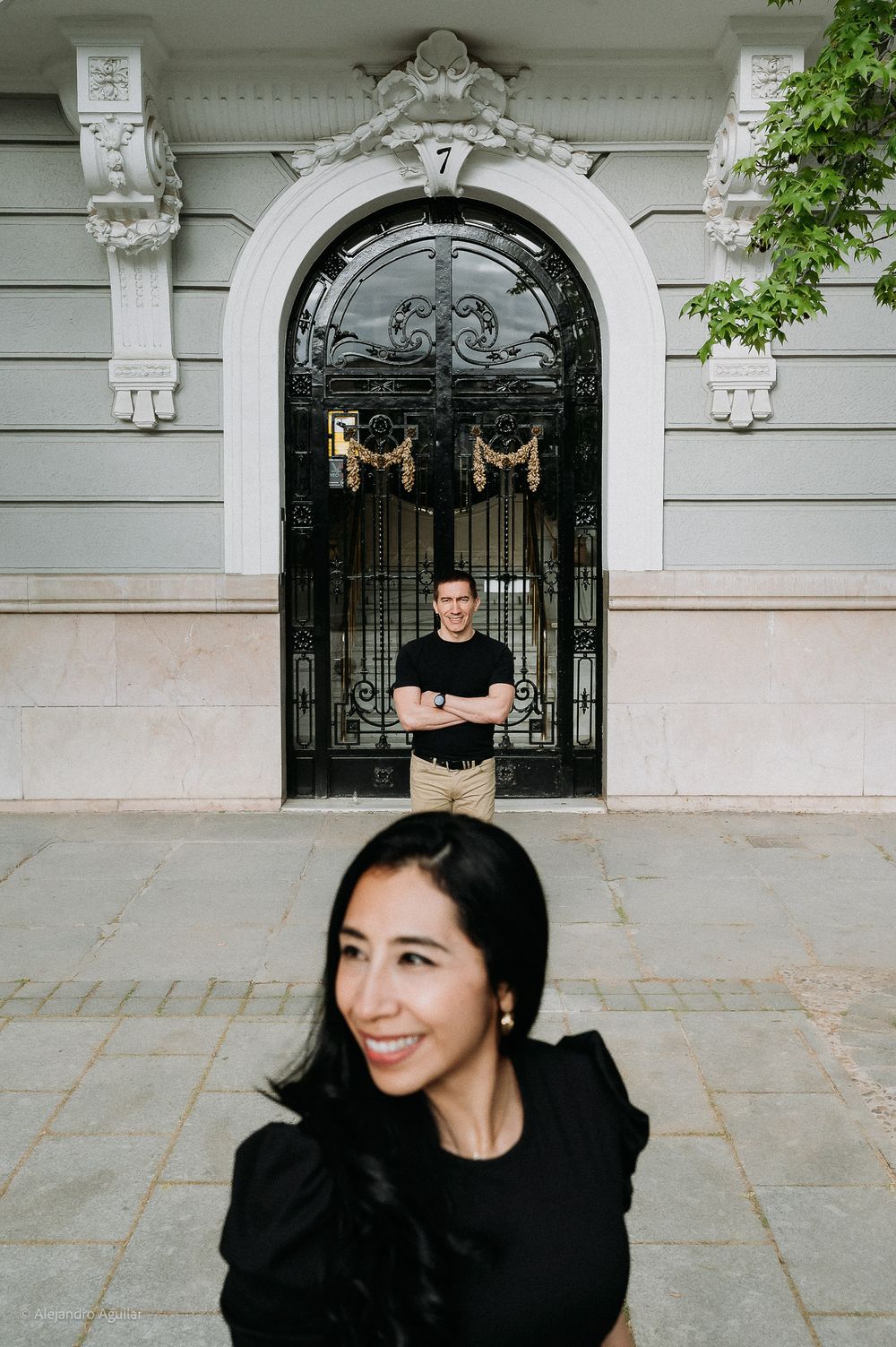 Playful couple posing at different depths before an ornate arched iron gate.
