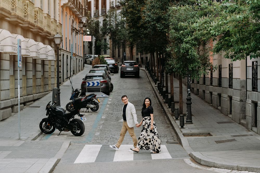 Couple crossing a quiet Madrid street lined with parked cars and trees.