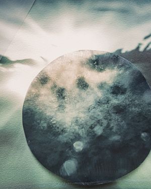 A circular piece of dark, weathered metal with abstract blue-gray tones rests on a light textured surface.