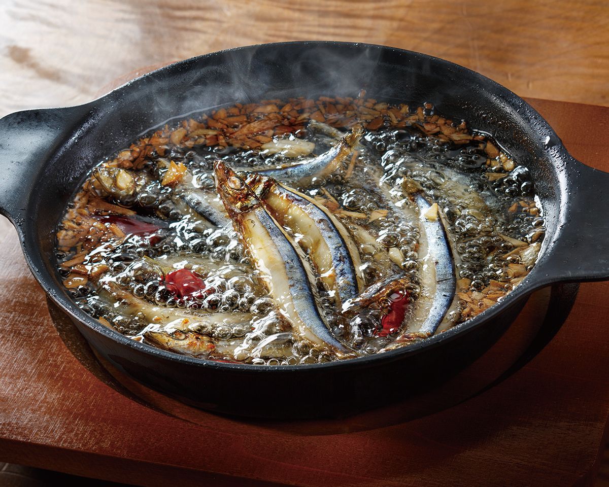 Sizzling anchovies with chili peppers cooking in oil in a cast iron skillet on a wooden surface.