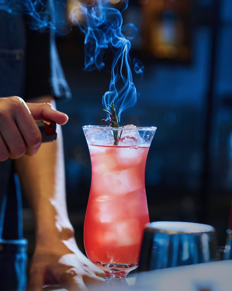 A bartender ignites a rosemary sprig on a red cocktail in a hurricane glass, creating dramatic blue smoke.