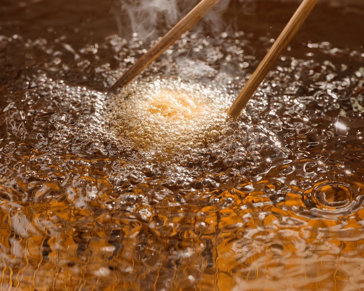 Food frying in hot bubbling oil with chopsticks stirring, steam rising from golden crispy batter in a deep fryer.