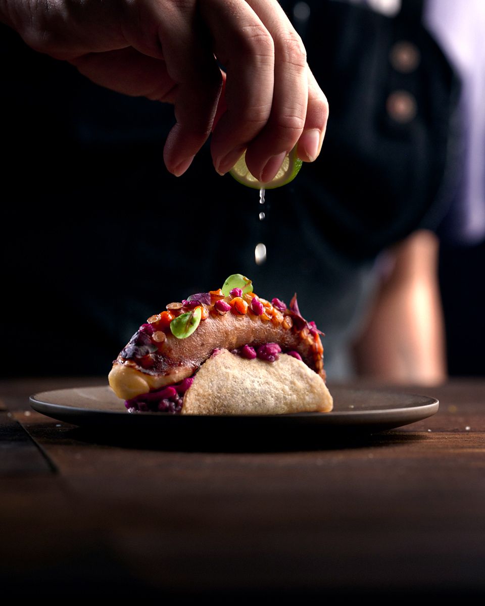 Chef squeezing fresh lime juice over a gourmet taco topped with colorful garnishes on a dark plate.
