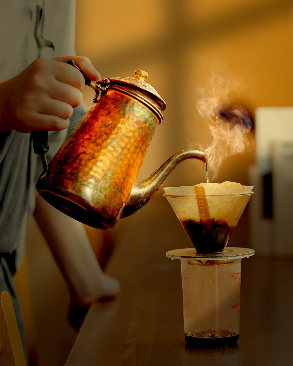 Person pouring hot water from a copper gooseneck kettle into a pour-over coffee dripper with steam rising.