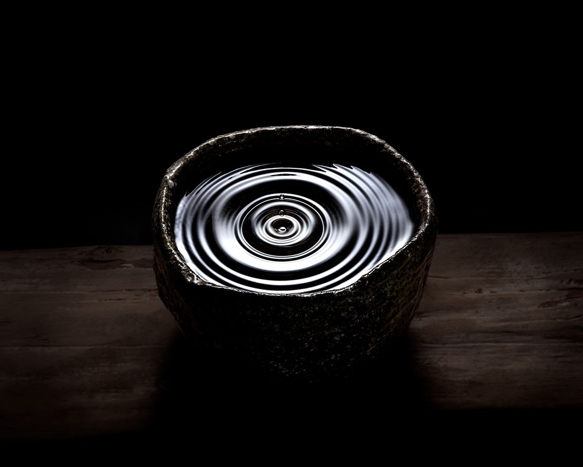 Dark ceramic bowl filled with rippling water creating concentric circles, set on a wooden surface against a black background.