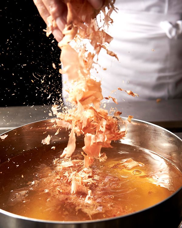 Chef adding bonito flakes to a steaming golden broth in a large stainless steel bowl.