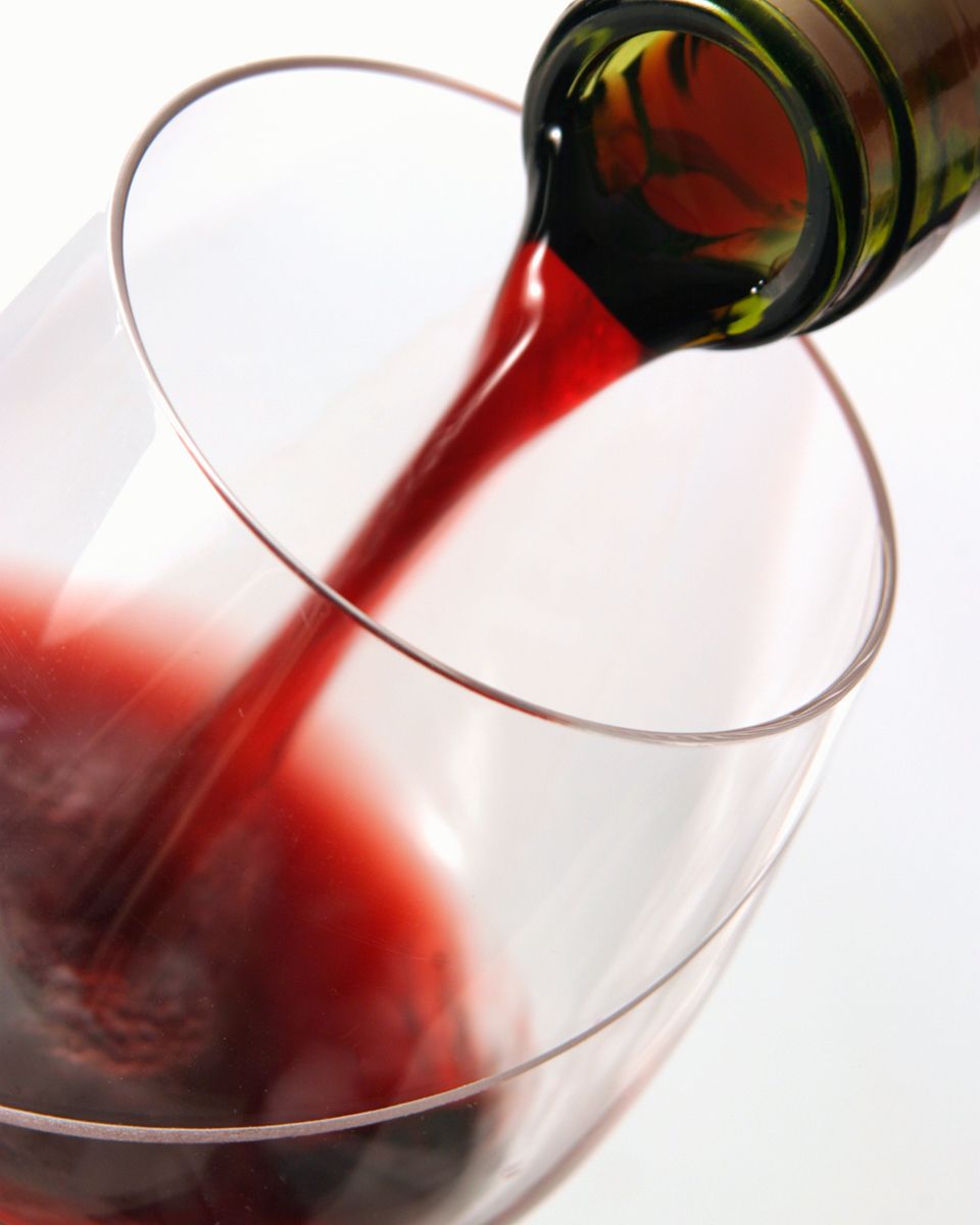 Red wine being poured from a green bottle into a large clear wine glass against a white background.