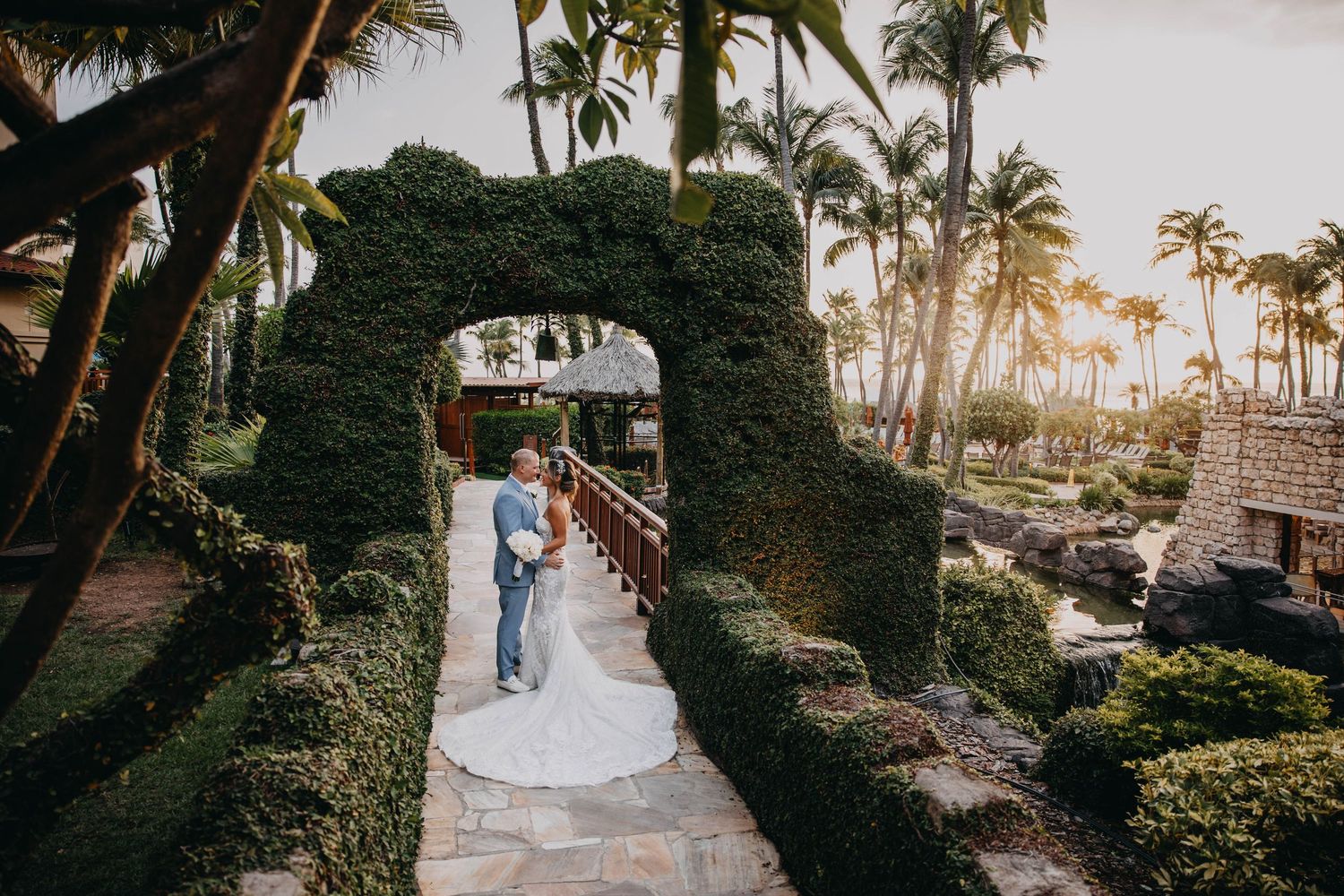 Bride and groom embrace under ivy-covered arch at Hyatt resort wedding venue with palm trees at sunset.