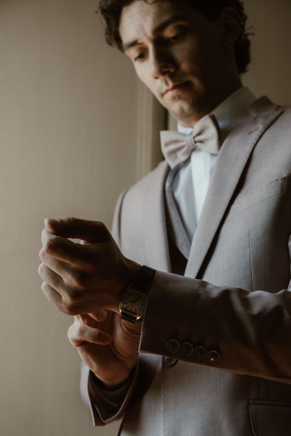Groom in gray suit and bow tie adjusting his gold watch while getting ready for wedding ceremony.