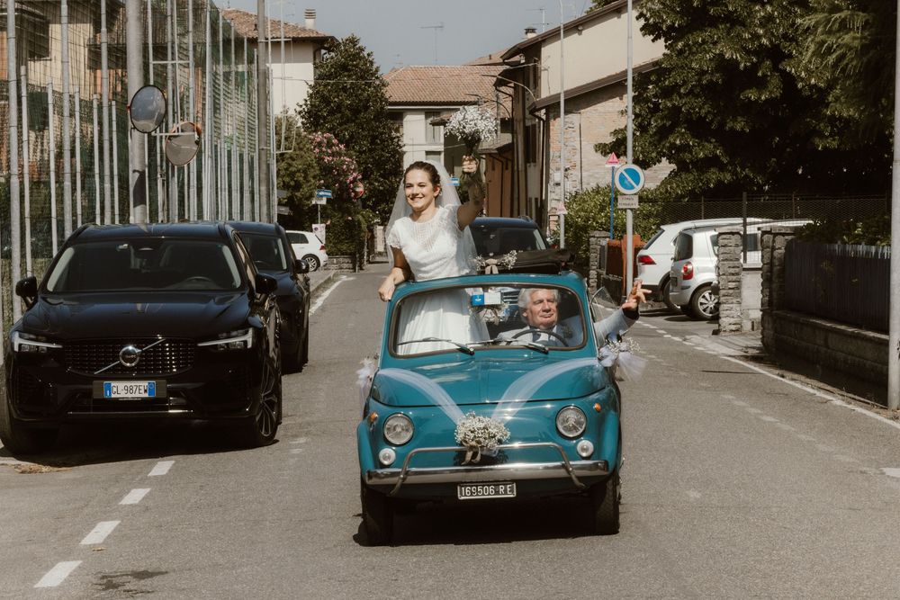 Bride standing in a classic blue Fiat 500 convertible driving through an Italian street on her wedding day.