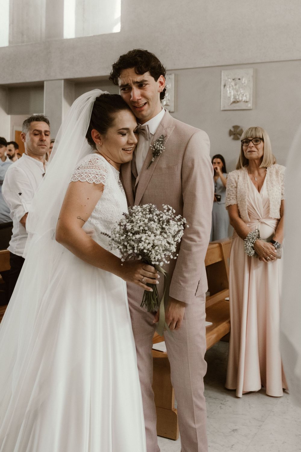 Bride in white lace gown holding baby's breath bouquet stands with groom in blush suit at church wedding ceremony.