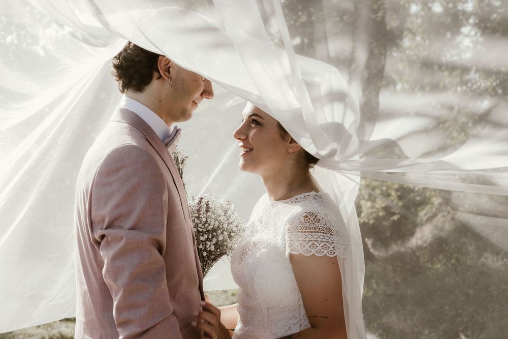 Bride and groom smiling under flowing white veil, groom in pink suit, bride in lace dress holding bouquet.
