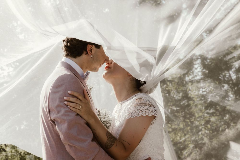 Bride and groom share a romantic kiss under a flowing white veil outdoors on their wedding day.