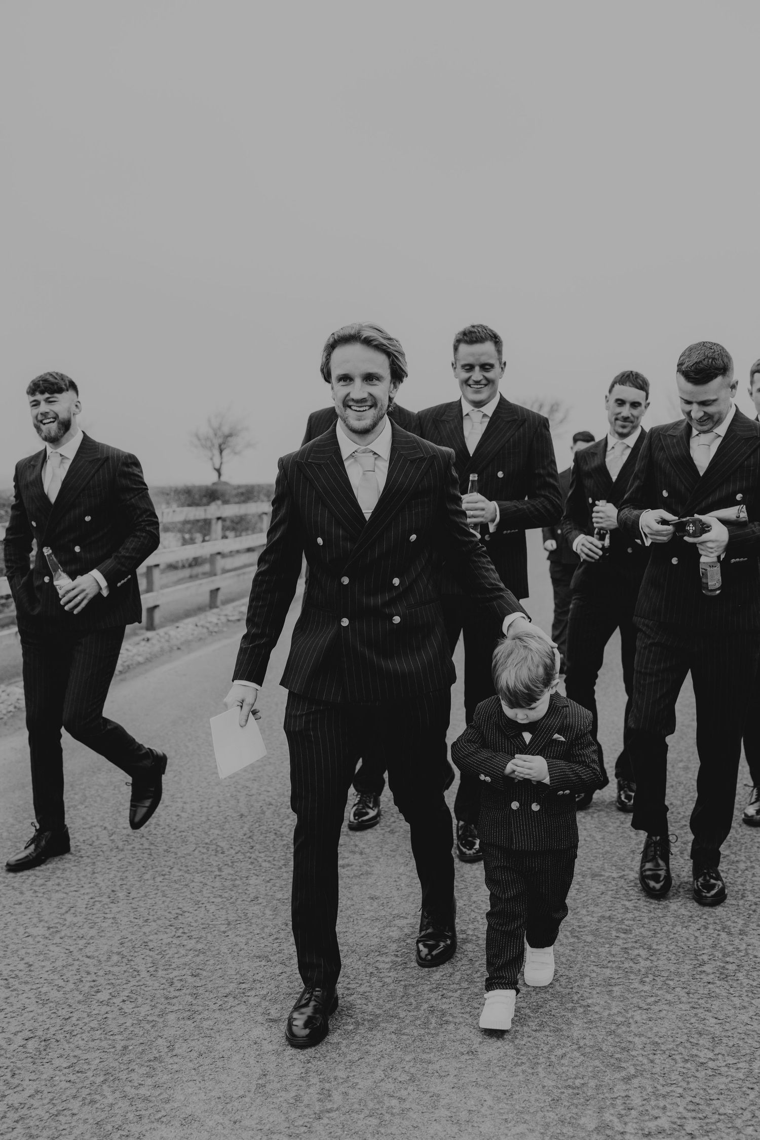Black and white photo of groom and groomsmen in suits walking confidently on a road at a wedding.