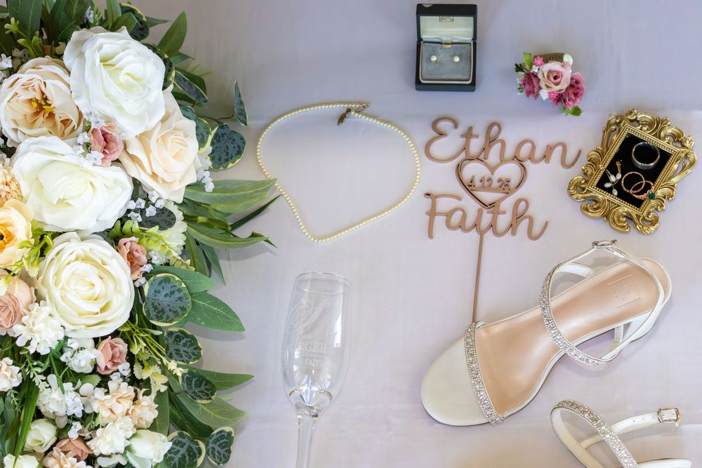 Wedding flat lay featuring floral bouquet, champagne flute, silver heels, jewelry, and a wooden Ethan and Faith cake topper.