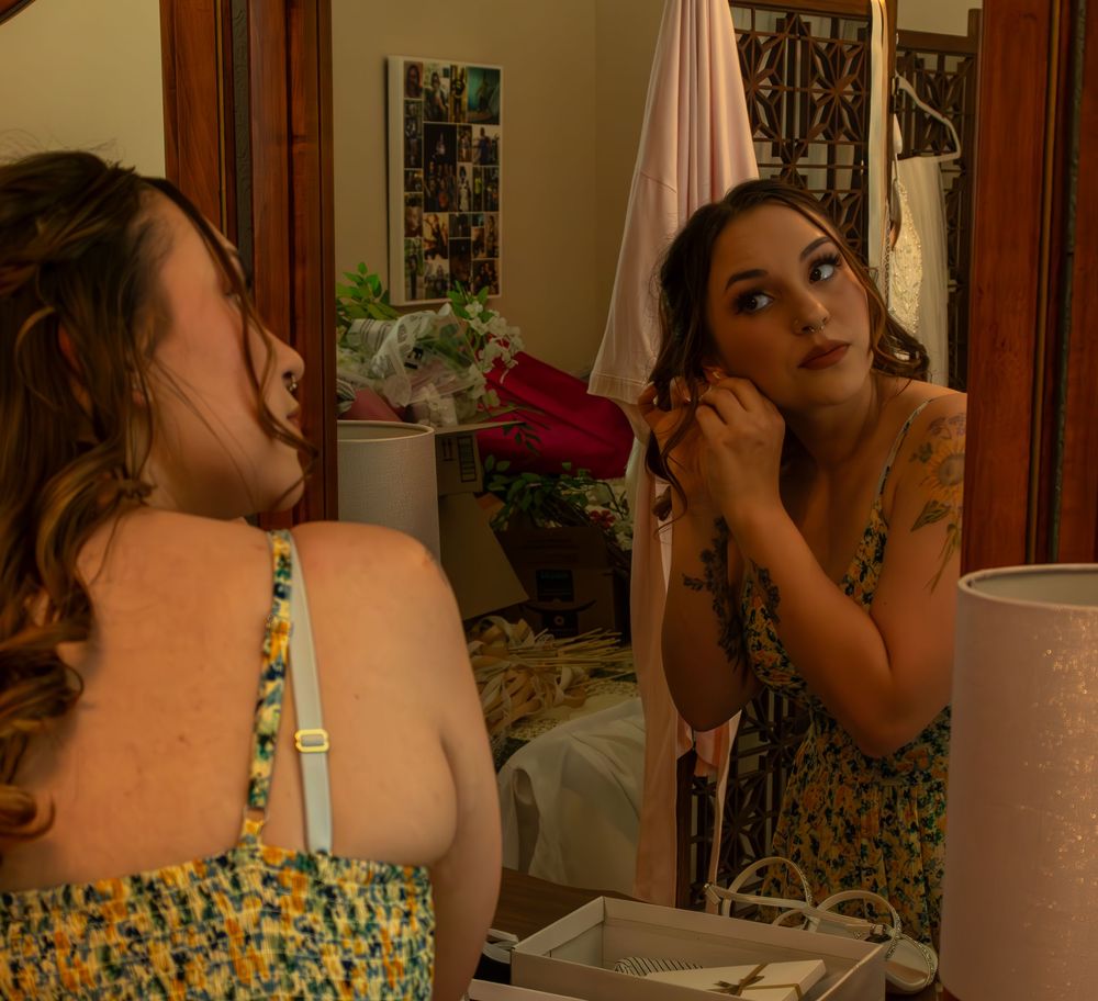 Young woman in floral dress applying earrings while looking in a bedroom mirror.