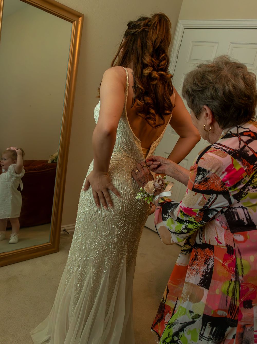 Elderly woman helping bride fasten her beaded wedding gown from behind, with mirror reflection visible.