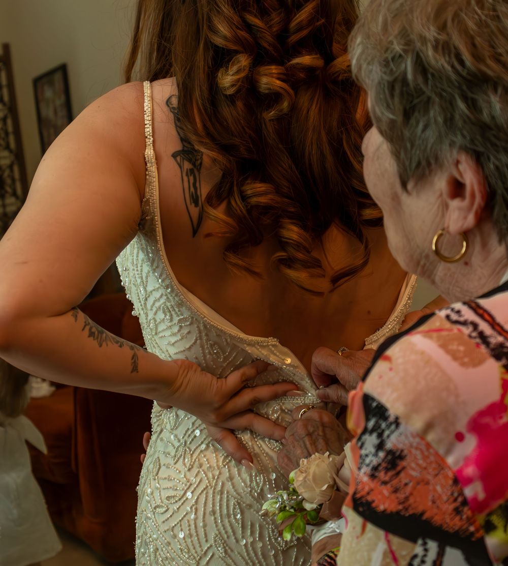Elderly woman helping bride fasten her beaded wedding gown from behind, showing intricate back detail.
