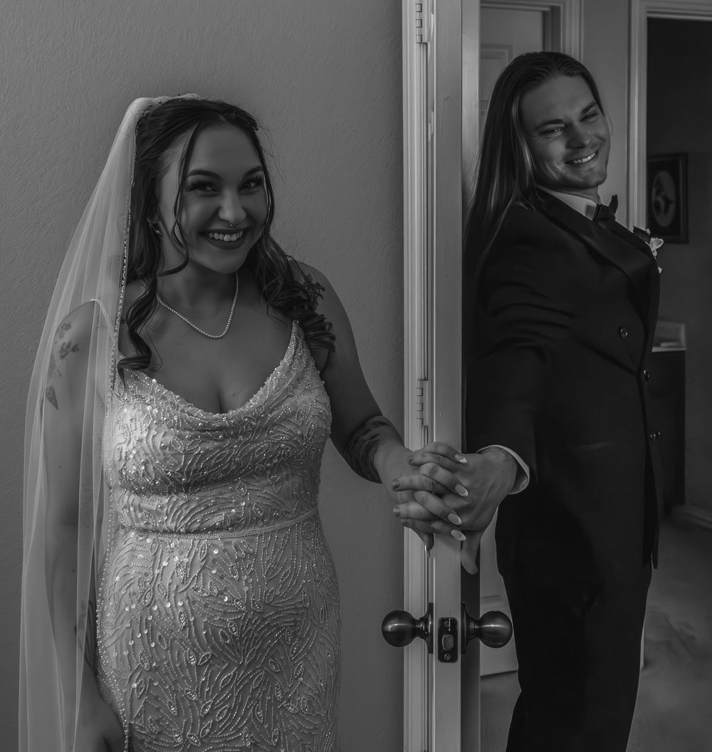Black and white photo of a bride in a beaded gown and veil holding hands with a groom in a suit by a door.
