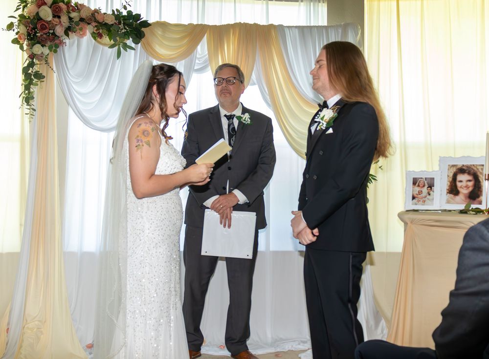 Bride reading vows to partner during indoor wedding ceremony with officiant and floral arch backdrop.